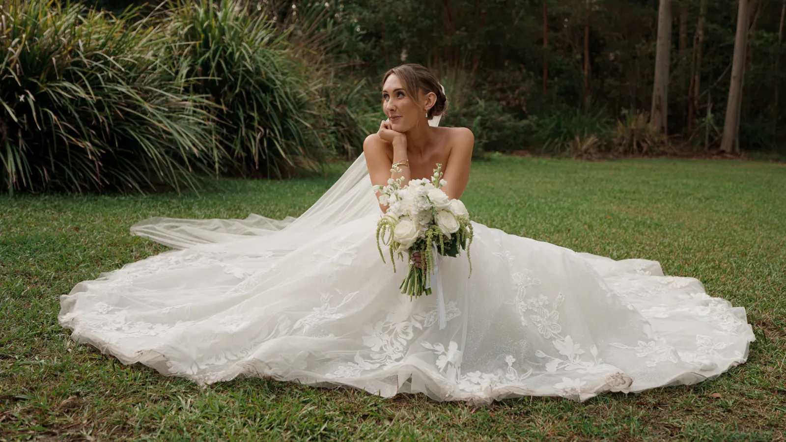 Shannon seated on the lawn in her wedding gown, the lace skirt and veil spread around her, her bouquet in hand